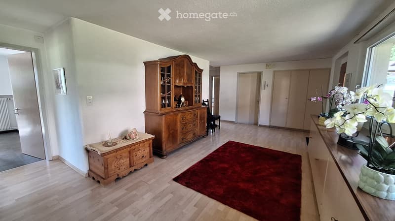 A well-lit hallway with wooden flooring, a red rug, a wooden cabinet, a small table with a decorative item, and a vase with white flowers on a console table.