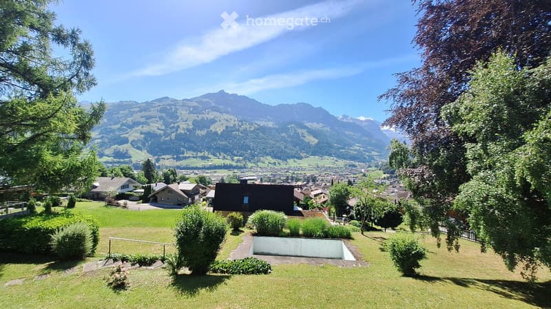 Garden with a swimming pool, surrounded by greenery, overlooking a town and mountain range.