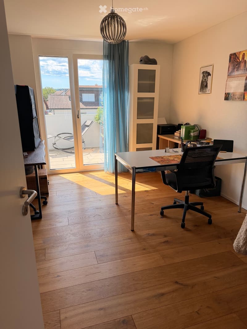 Brightly lit room with wooden flooring, a large window with blue curtains, a desk with a chair, a cabinet, and a TV on a stand.