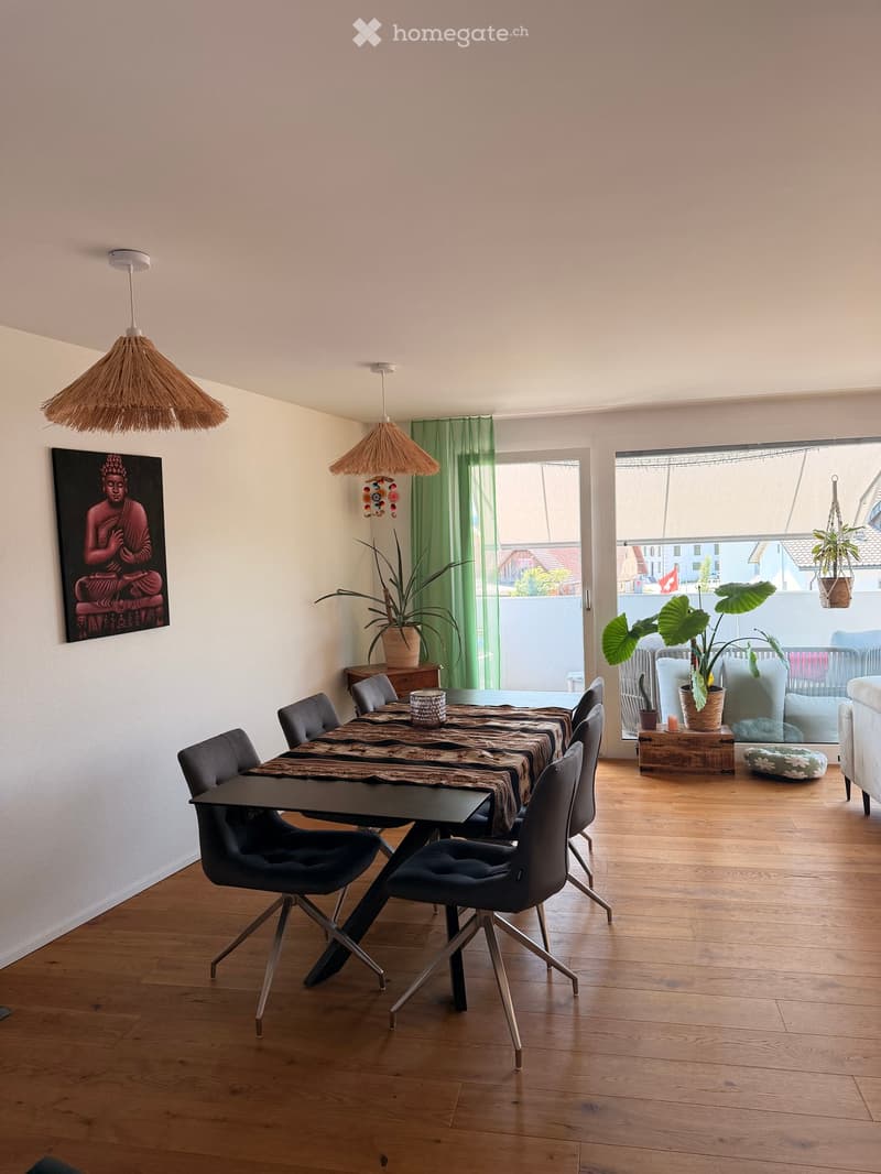 Dining area with a black dining table, six chairs, pendant lights, a painting, and plants. The room has wooden flooring and large windows with green curtains.