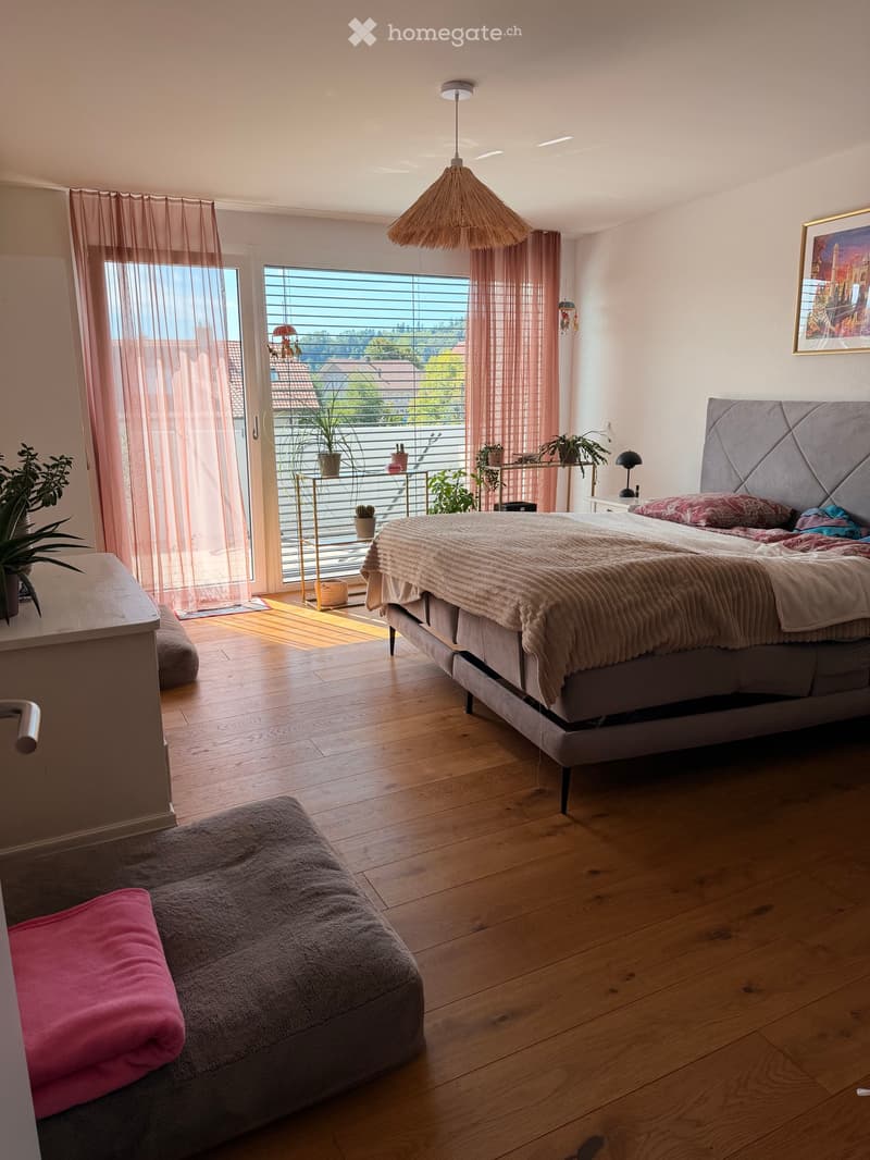 Bright bedroom with wooden flooring, large bed with headboard, nightstand, chair, small table, potted plants, and a view of the balcony.