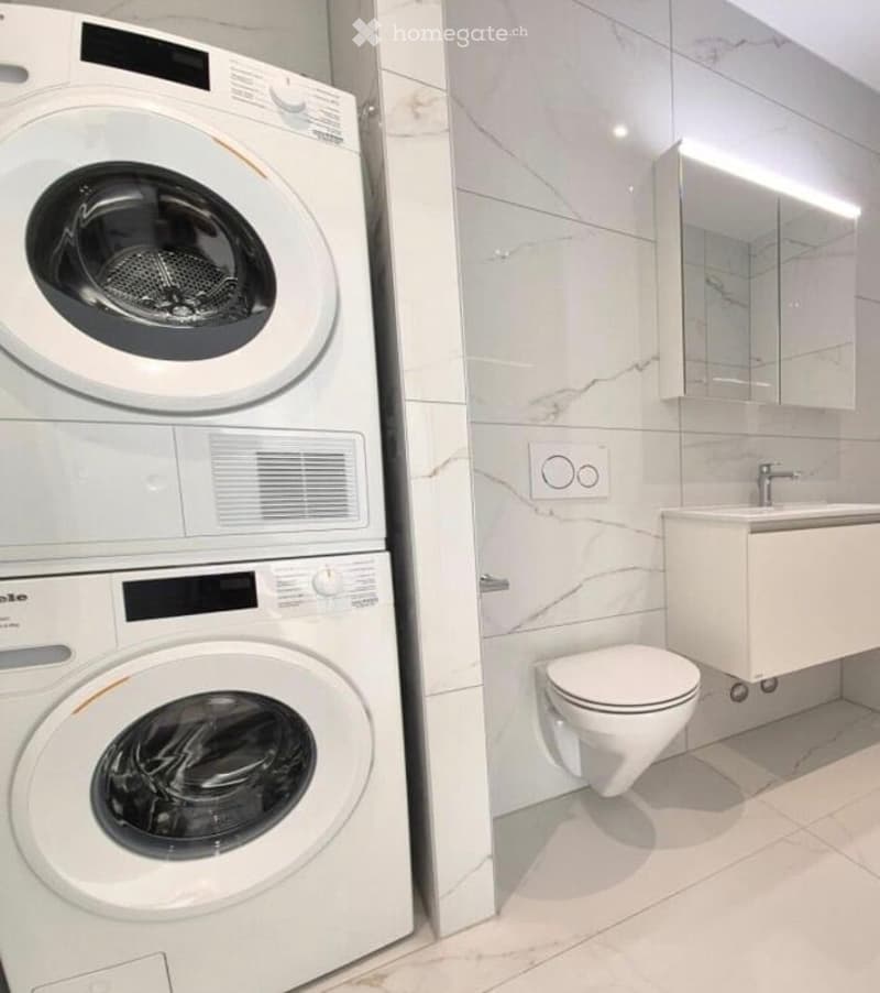 Modern bathroom with white marble tiles, a toilet, a sink with a mirror, and a stacked washing machine/dryer unit.