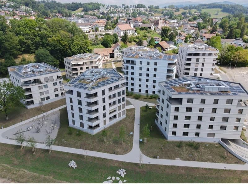 Aerial view of a modern residential complex with multiple white apartment buildings featuring solar panels on the rooftops, surrounded by greenery and pathways.