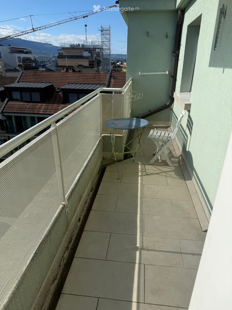 Balcony with tiled floor, metal railing, small round table, and folding chair, overlooking cityscape with rooftops and mountains.