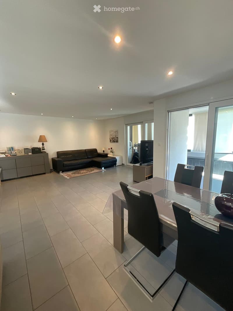 Modern living and dining area with a black leather sofa, glass dining table with chairs, a side cabinet, and a lamp. The space features large windows and a sliding glass door leading to a balcony.
