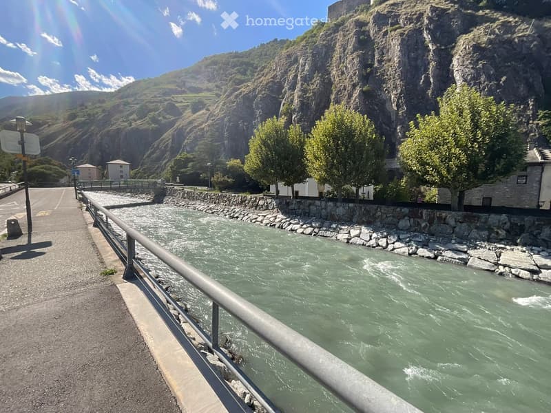A scenic riverside view with a flowing river, rocky banks, trees, and distant mountains under a clear blue sky.