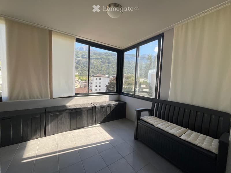 Balcony with large windows offering a scenic mountain view, featuring a wooden bench with cushions and several storage boxes.
