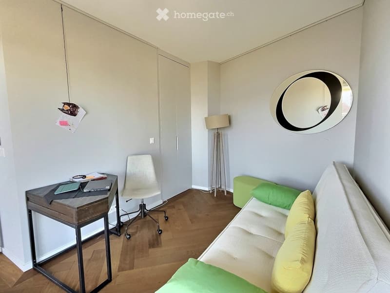 A modern, well-lit room with a sofa, a chair, a small table with a laptop, a floor lamp, a round mirror, and a green ottoman.
