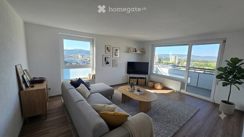 Bright living room with large windows offering a scenic view, featuring a grey sectional sofa, wooden coffee table, TV, bookshelf, and decorative plant.