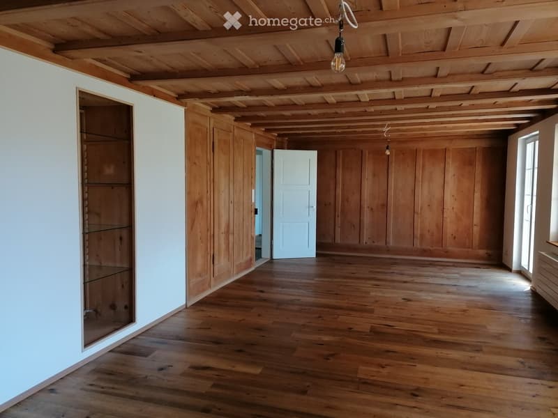 Empty room with wooden floors, white walls, exposed wooden ceiling beams, and a glass-fronted cabinet.