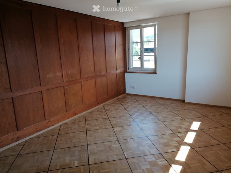 Empty room with wooden panel walls and herringbone-patterned wooden flooring, featuring a single window with white frame and a visible electrical outlet on the wall.