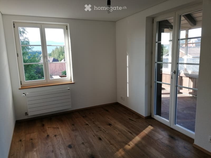 Empty room with wooden flooring, white walls, large window with view, sliding glass door leading to balcony, radiator, and ceiling light fixture.
