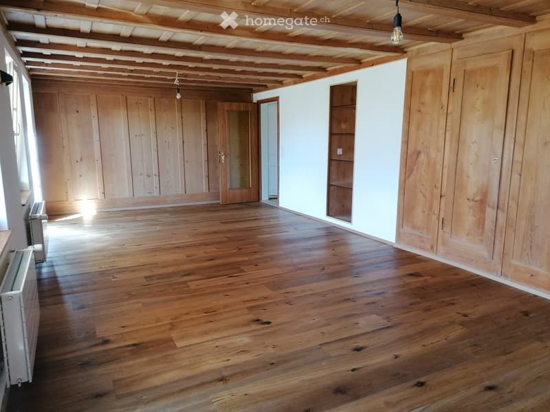 Empty room with wooden floors, exposed wooden beams on the ceiling, and large wooden doors. Natural light streams in from the left side.