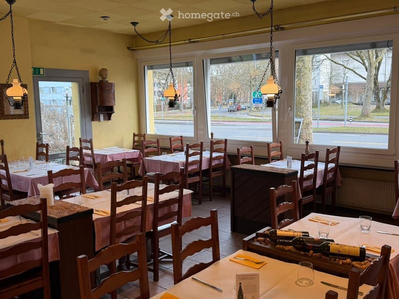 Restaurant interior with multiple tables and chairs, each set with wine bottles, menus, and cutlery. Large windows provide a view of the street outside.