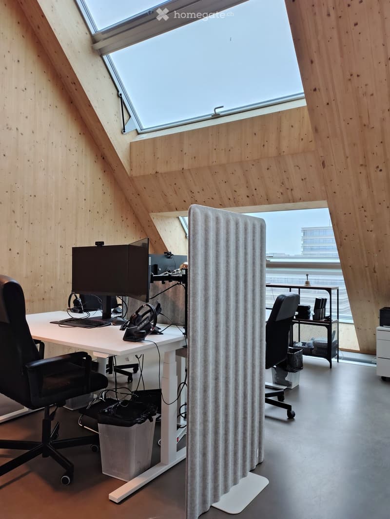 Modern office space with wooden walls, large skylight windows, and a sleek desk setup featuring dual monitors, ergonomic chairs, and a screen divider for privacy.