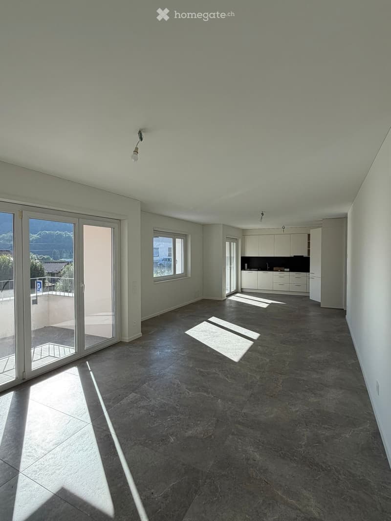 Empty room with grey tiled floor, white walls, large sliding glass doors leading to a balcony, and a small kitchen area with white cabinets and a countertop.
