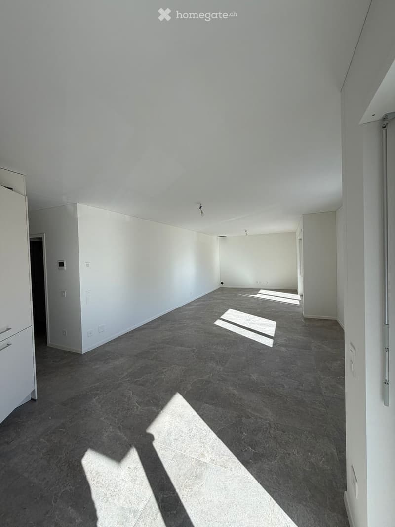Empty room with white walls, dark grey tiled floor, and natural light streaming in from a window. No furniture or equipment visible.