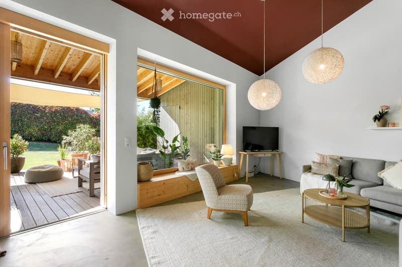 Modern living room with large windows, grey sofa, coffee table, armchair, TV, and decorative items.