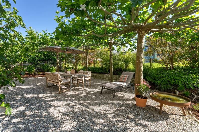 A serene garden area with a gravel-covered ground, featuring a wooden bench, a lounge chair, a small table with chairs, a potted plant, and a decorative water fountain. The area is shaded by large trees and surrounded by lush greenery.
