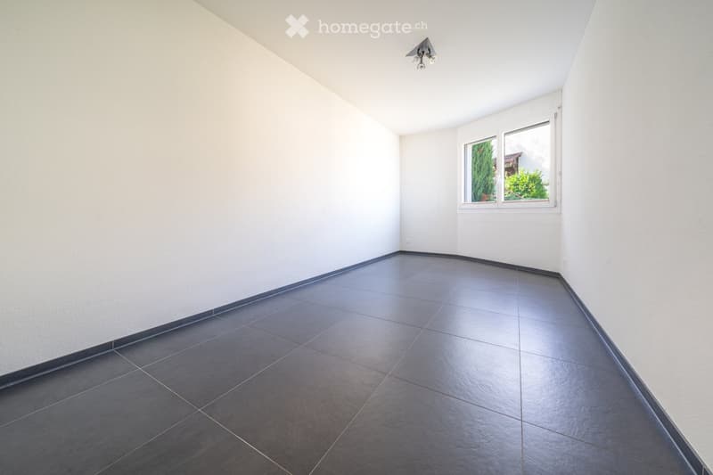 Empty room with white walls, dark tiled floor, and a single window with white frame. No furniture or equipment present.