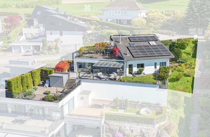 Aerial view of a modern, multi-story house with a flat roof, solar panels, and a spacious garden area. The house features large windows, a balcony, and a patio with outdoor furniture. The surrounding area includes other houses, greenery, and a driveway wi
