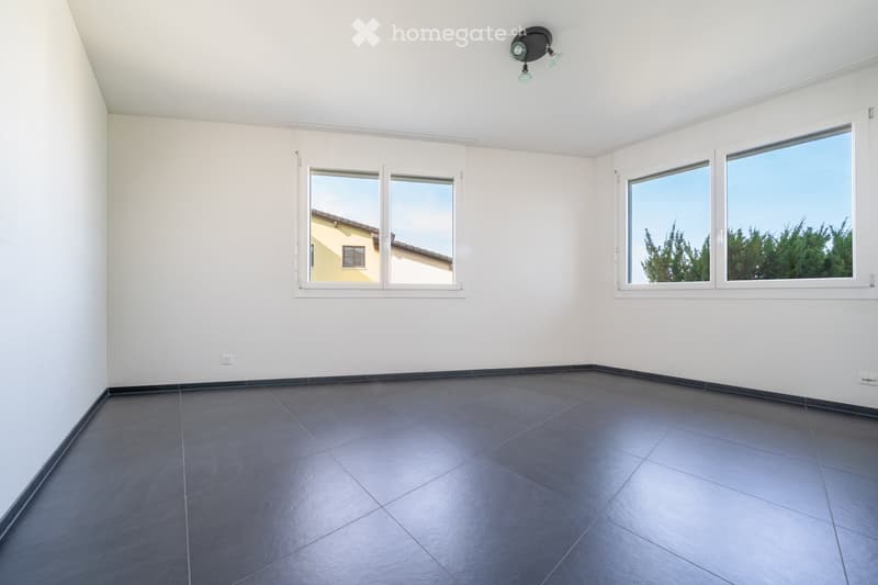 Empty room with white walls, dark tiled floor, and large windows offering views of a neighboring building and greenery.