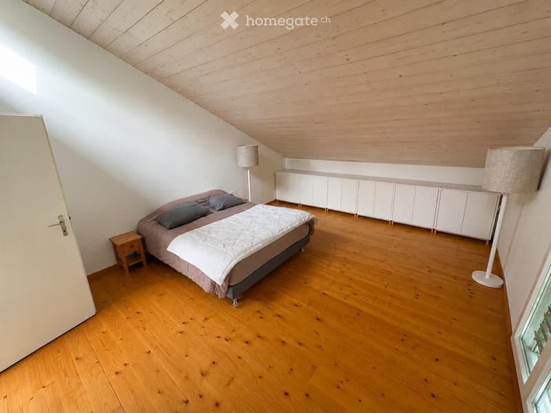 Bright bedroom with wooden flooring, slanted wooden ceiling, bed with pillows and duvet, nightstand, white cabinet, and floor lamps.
