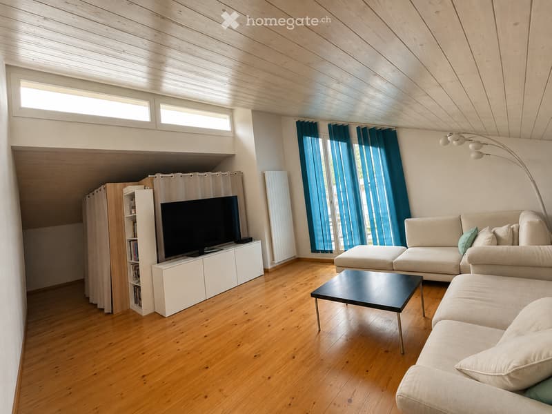 Modern living room with wooden floors, white walls, and large windows with blue curtains. Features a beige sectional sofa, black coffee table, flat-screen TV on a white stand, bookshelf, and a floor lamp.