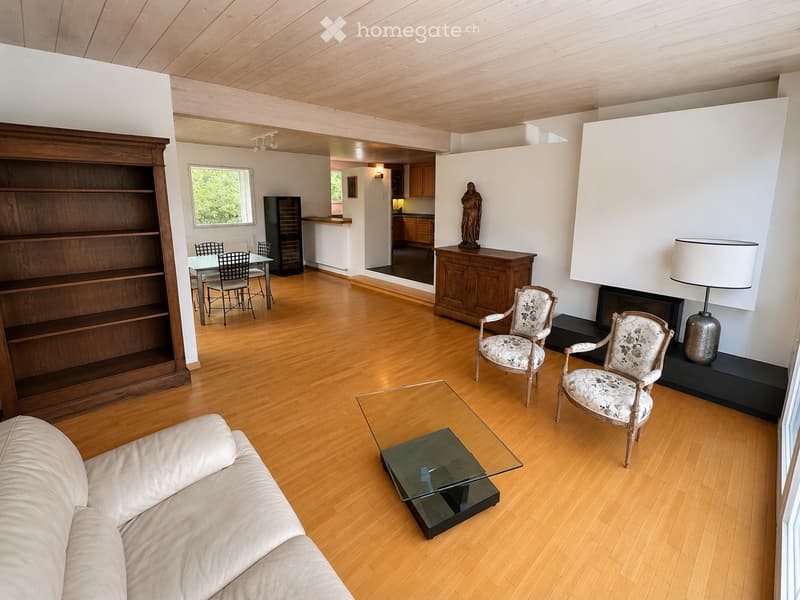 Spacious living room with wooden flooring, a cream-colored sofa, glass coffee table, two ornate armchairs, a wooden cabinet, a fireplace with a lamp, and a dining area with a table and chairs.
