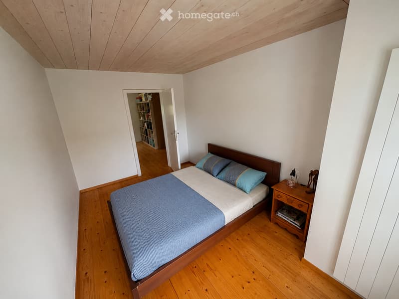 Cozy bedroom with wooden floor, slanted wooden ceiling, bed with blue bedding, nightstand with lamp, and open door leading to a bookshelf.