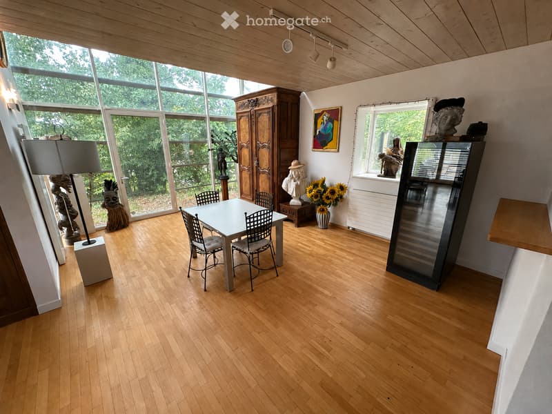 Bright room with wooden flooring, large windows, a dining table with chairs, a cabinet, a vase with sunflowers, a painting, and sculptures.