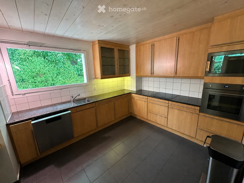 Modern kitchen with wooden cabinets, stainless steel appliances, and a large window overlooking greenery.