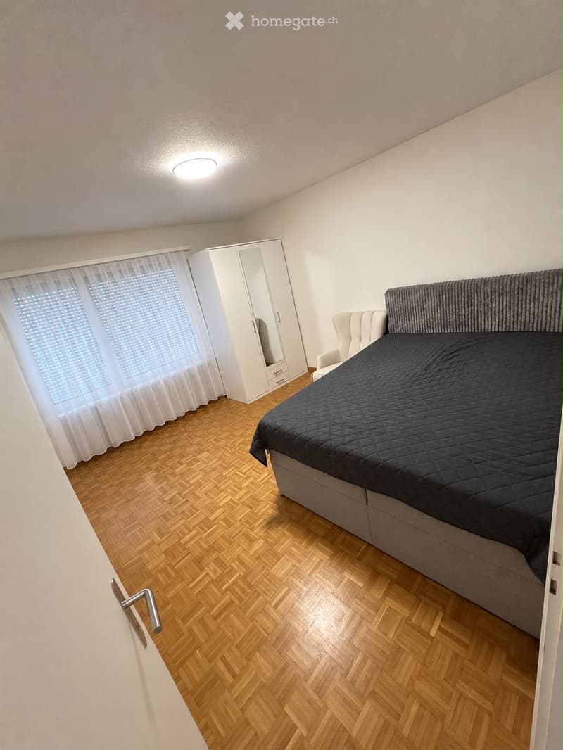 Modern bedroom with wooden herringbone flooring, a large bed with dark bedding, a white wardrobe with mirrored doors, and a white armchair near the window.