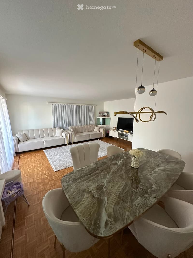 Modern living and dining area with a white sofa, grey dining table, chairs, and a television. The room features wooden flooring, a large window with curtains, and modern lighting fixtures.