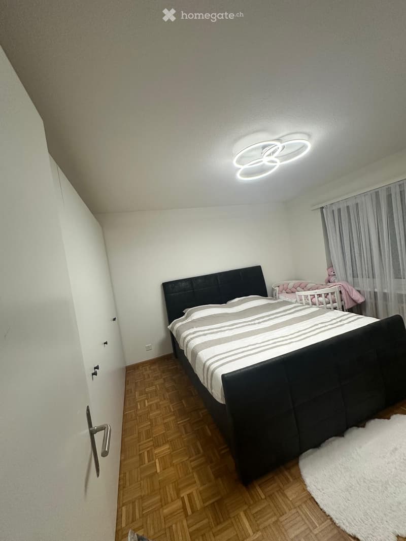 Modern bedroom with a large bed, nightstand, and a small crib. Features wooden flooring, white walls, and a contemporary ceiling light fixture.