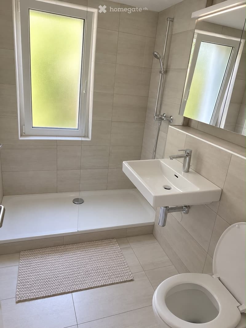 Modern bathroom with a bathtub, sink, toilet, and showerhead. Features a large window with frosted glass, beige tiles, and a small textured rug.