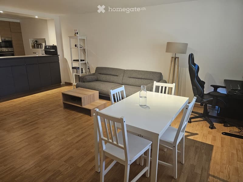 Modern living and dining area with a grey sofa, wooden coffee table, white dining table with chairs, floor lamp, and a small shelf with items. The space features a kitchen area with dark cabinets and appliances.