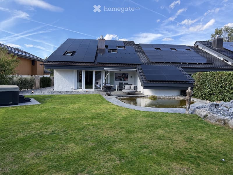 A modern house with solar panels on the roof, surrounded by a well-maintained lawn, a small pond, a patio area with outdoor furniture, and a hot tub.