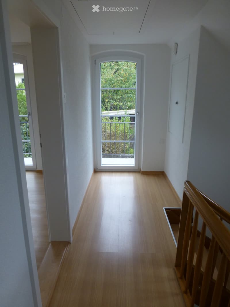 Bright hallway with wooden flooring, white walls, and a staircase leading upwards. A large window at the end provides natural light and a view of greenery outside.