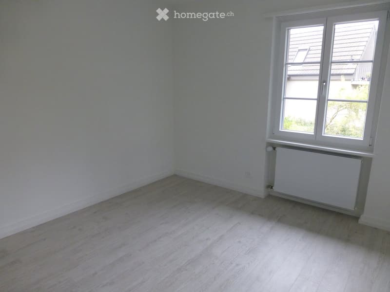 Empty room with light-colored wooden flooring and white walls, featuring a large window with white frames and a radiator below it.