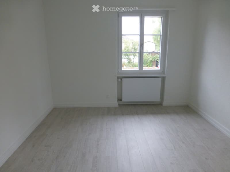 Empty room with light wooden flooring, white walls, and a single window with white frame. A radiator is visible below the window.