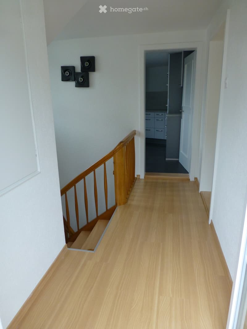 A clean, well-lit hallway with light wooden flooring and white walls. A wooden staircase with a handrail is visible on the left, and a doorway leads to another room on the right.