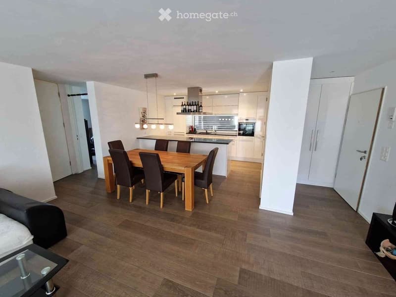 Dining room with wooden dining table, black chairs, modern kitchen with island, wooden floor, white walls, white cupboards, dining table, lights, microwave, and kitchen cabinets.