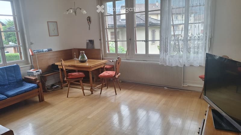 A cozy living room with wooden flooring, a blue sofa, a wooden dining table with chairs, a television, and a vase on the table. Large windows with white curtains let in natural light.