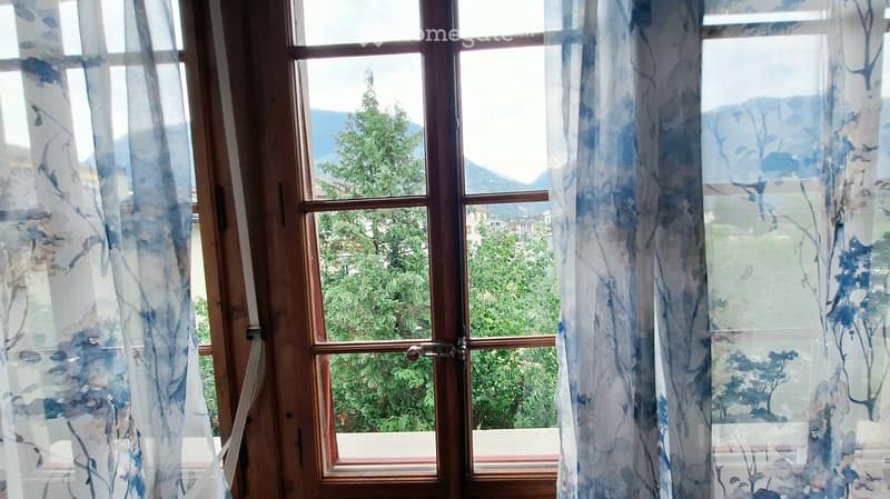 An unfurnished room with large windows covered by sheer curtains, offering a scenic view of greenery and mountains.
