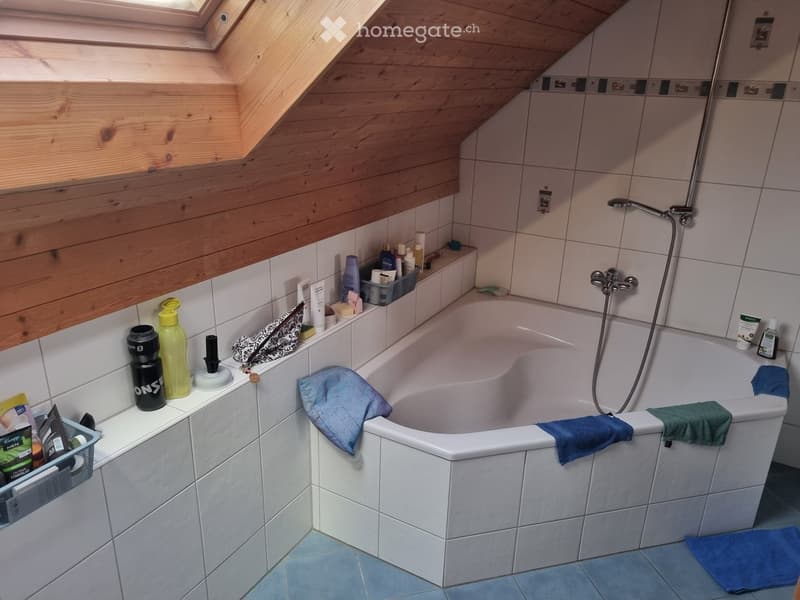 Bathroom with a white bathtub, tiled walls, wooden slanted ceiling, and various toiletries on the edge of the tub.