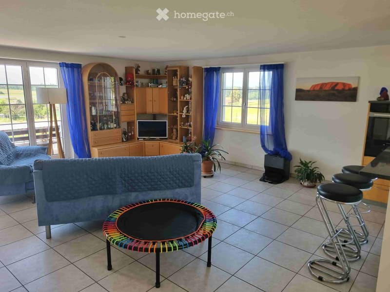 Bright living room with large windows, blue curtains, a blue sofa, a colorful round coffee table, bar stools, a wooden cabinet, and a television. The room features tiled flooring and potted plants.