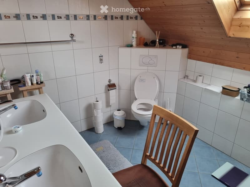 Modern bathroom with white tiles, a bathtub, toilet, sink, wooden chair, and various toiletries.