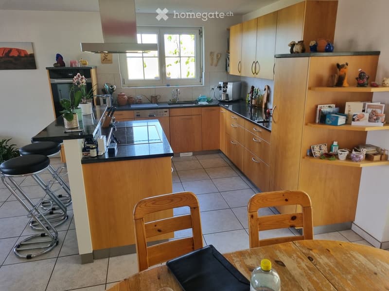 Bright kitchen with wooden cabinets, black countertops, and a central island with bar stools. Includes a sink, oven, and various kitchen items on shelves and counters.