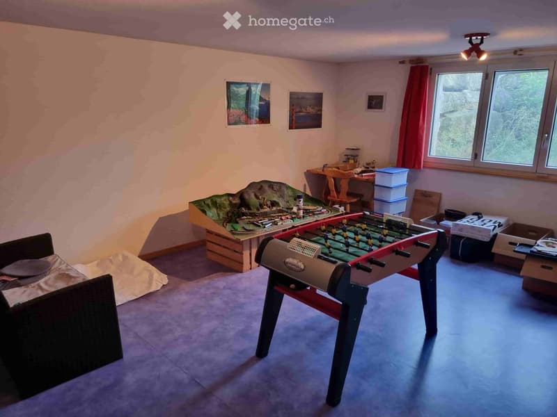 A cozy room with a sofa, futon, train set, foosball table, and storage boxes. The room has large windows with red curtains and is well-lit with ceiling spotlights.